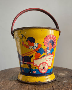 A Vintage Child's Tin Beach Bucket in sunny bright colours. The delightful scene on the side shows a donkey pulling along a lad giving a lift to a young lady with her parasol. These tin buckets are becoming increasingly rare and are very collectable - SHOP NOW - www.intovintage.co.uk