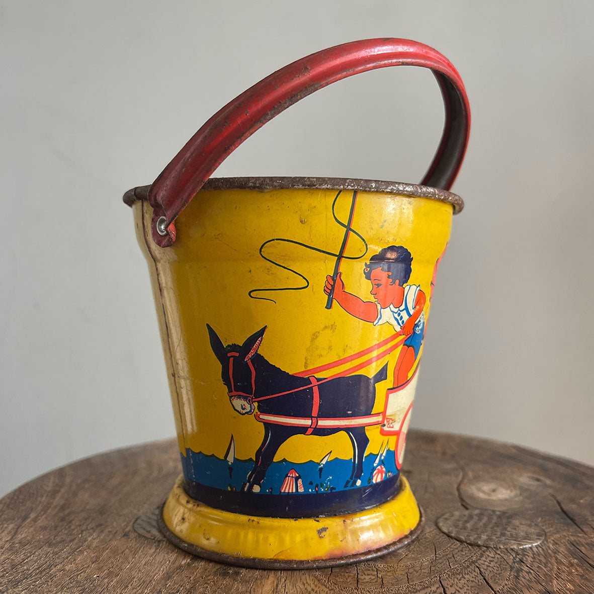 A Vintage Child's Tin Beach Bucket in sunny bright colours. The delightful scene on the side shows a donkey pulling along a lad giving a lift to a young lady with her parasol. These tin buckets are becoming increasingly rare and are very collectable - SHOP NOW - www.intovintage.co.uk