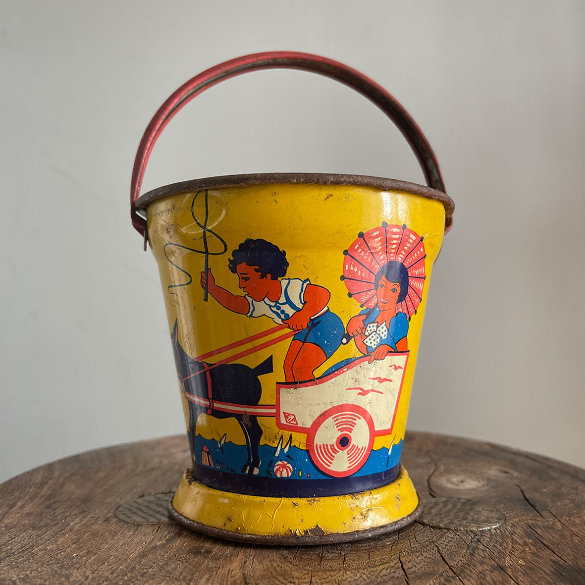 A Vintage Child's Tin Beach Bucket in sunny bright colours. The delightful scene on the side shows a donkey pulling along a lad giving a lift to a young lady with her parasol. These tin buckets are becoming increasingly rare and are very collectable - SHOP NOW - www.intovintage.co.uk